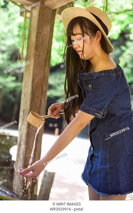 Young woman wearing blue dress and hat using bamboo water hand washing basins at Shinto Sakurai Shrine, Fukuoka, Japan