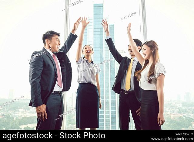 Group of smiling businesspeople standing near the window raising their hands