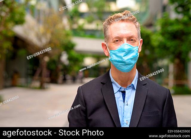 Portrait of mature businessman with mask for protection from corona virus outbreak in the city with nature outdoors