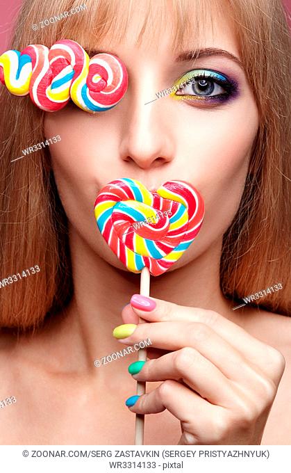 Beauty portrait of young blonde woman on pink background. Female with candy lollipop on stick in hands. Girl has finger nails with bright yellow, green