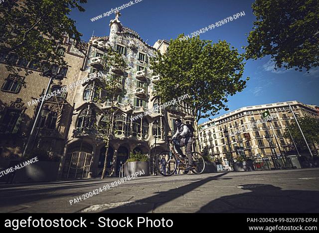 24 April 2020, Spain, Barcelona: A man wearing a face mask against the coronavirus rides his bike through a deserted street past the ""Casa Batllo"" of the...