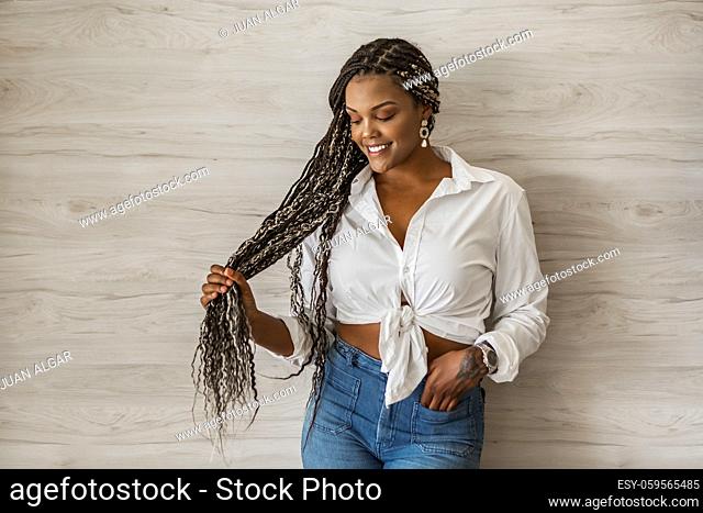 Portrait of a cheerful African American adult woman holding her long hair. Adult woman with black and yellow braids posing on a wall background and looking down
