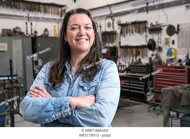 Woman with brown hair wearing Denim shirt standing in metal workshop, smiling at camera