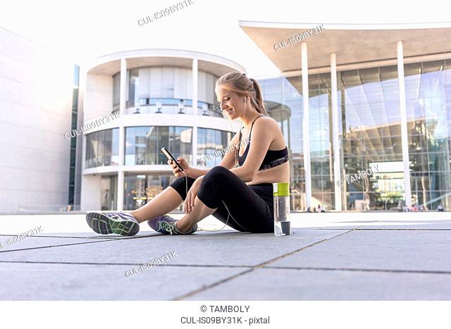 Young woman taking break from exercise and using smartphone in city, Berlin, Germany