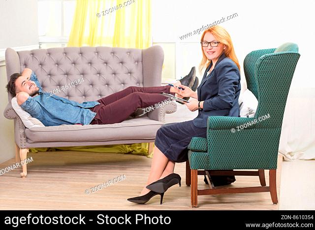 Patient man relaxing on a couch in front of a female psychiatrist with clipboard. View of sad man talking with therapist