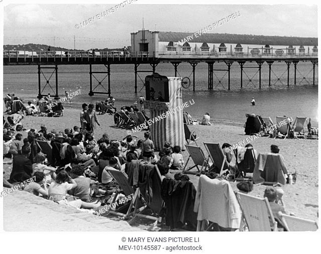 Rows of holidaymakers, mostly seated in deckchairs, watch the Punch and Judy show on the beach at Paignton, Devon, England