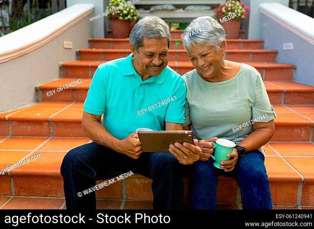 Smiling biracial senior couple holding coffee cups and using digital tablet while sitting on steps