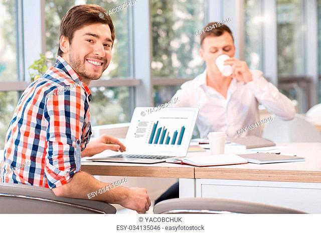 Get some rest. Cheerful delighted handsome man smiling working on the laptop and sitting at the table while his colleague drinking coffee in the background