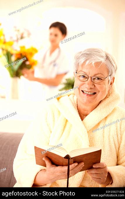 Portrait of happy elderly woman sitting on couch, reading book, nurse arranging flowers in background
