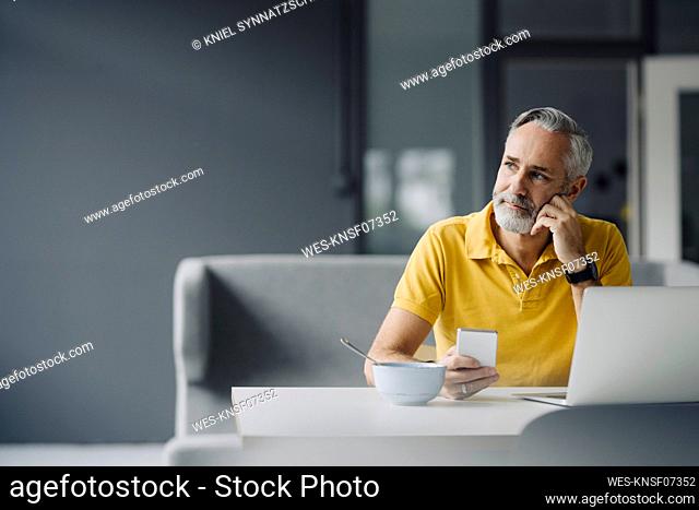 Portrait of mature man sitting at table with laptop and smartphone looking at distance
