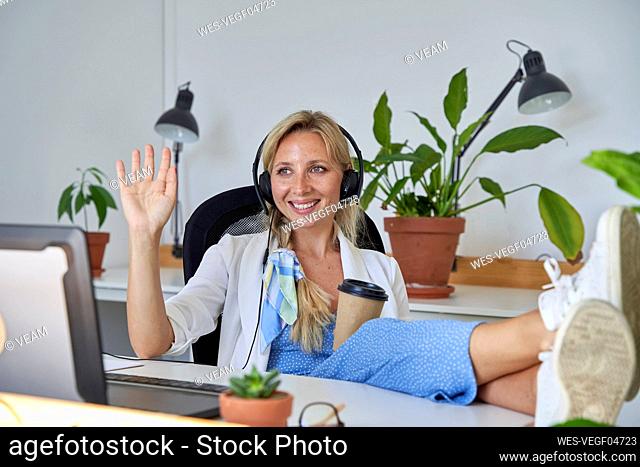 Smiling female customer service representative waving while talking during video call at office