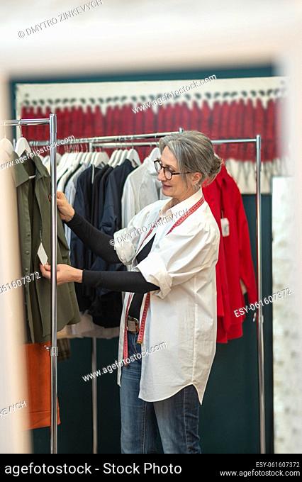 Good result. Middle age woman in glasses smiling looking with pleasure touching price tag on blouse standing near rack with clothes