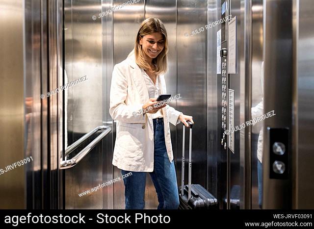 Smiling businesswoman using smart phone standing with suitcase in elevator