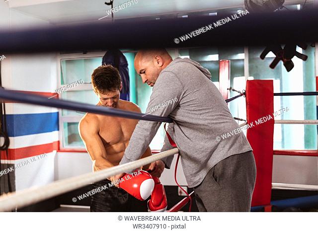 Side view close up of a young Caucasian male boxer by a boxing ring having his boxing gloves checked by a middle aged Caucasian male trainer