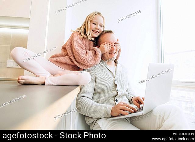 Happy daughter covering eyes of father sitting with laptop by window at home