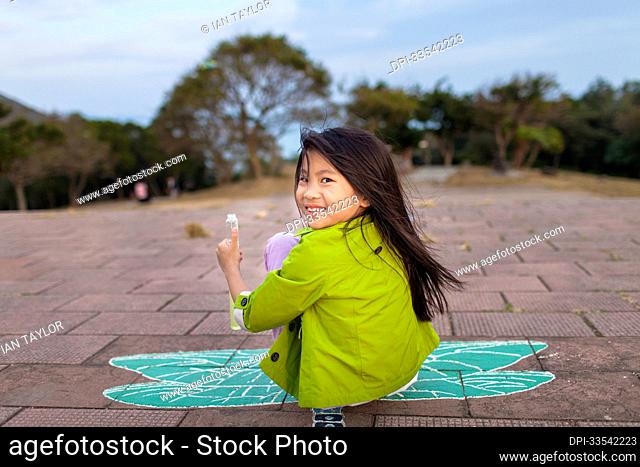 Young girl sits on the ground on a painted butterfly, looking back at the camera and giving a thumbs up; Hong Kong, China