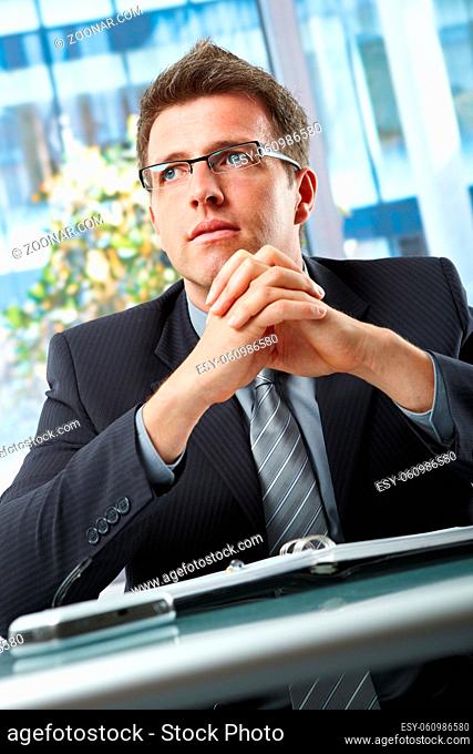 Confident businessman wearing suit and glasses looking aside with hands folded over oraniser in office