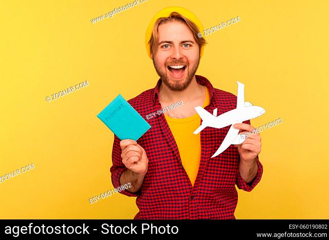 Positive hipster tourist guy in beanie hat, checkered shirt holding paper plane and passport, rejoicing tour abroad, visa free travelling by air transportation