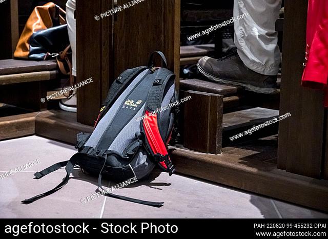 20 August 2022, Bavaria, Würzburg: A hiking backpack lies on the floor next to a pew where a pilgrim with hiking boots is sitting
