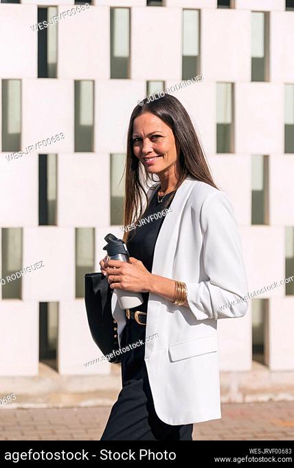 Smiling businesswoman with insulated drink container walking by wall
