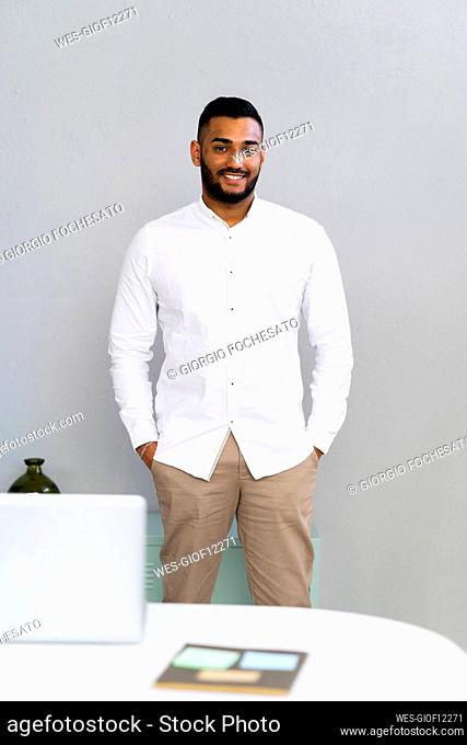 Young businessman with hands in pockets standing by wall in office