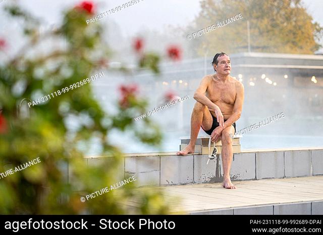 PRODUCTION - 17 November 2023, North Rhine-Westphalia, Oelde: Hans Ulrich Remfert sits on a starter block in the outdoor pool while steam rises from the...
