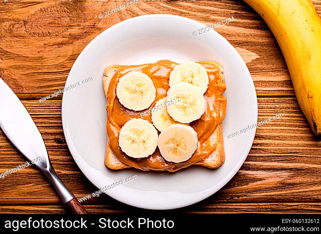 Toast with peanut butter and banana on white ceramic plate with knife on rustic wooden background, top view flat lay