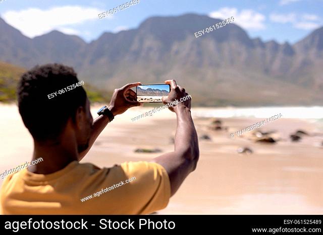 African american man taking photo of the mountains with smartphone on a beach by the sea