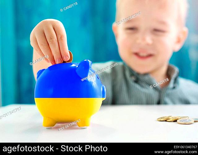 Child sitting at the table with money coins and biggybank in colour of Ukranian flag. Concept of the assistant, helping and donation for Ukraine