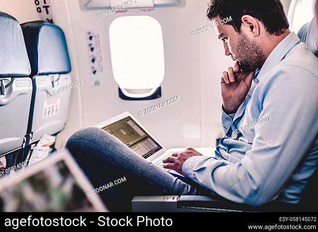 Casually dressed middle aged man working on laptop in aircraft cabin during his business travel. Shallow depth of field photo with focus on businessman eye