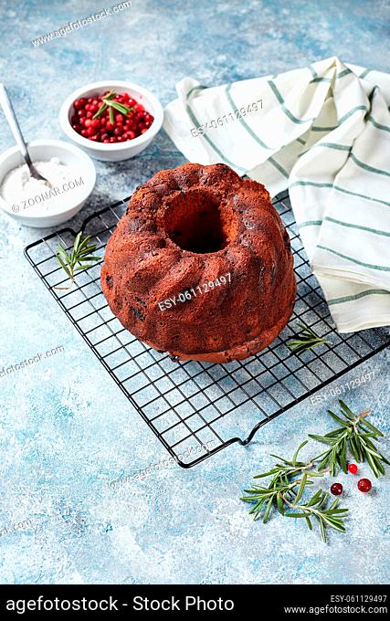 Chocolate bundt cake on a metal wire rack, powdered sugar and cranberries in bowls, prepared for decoration