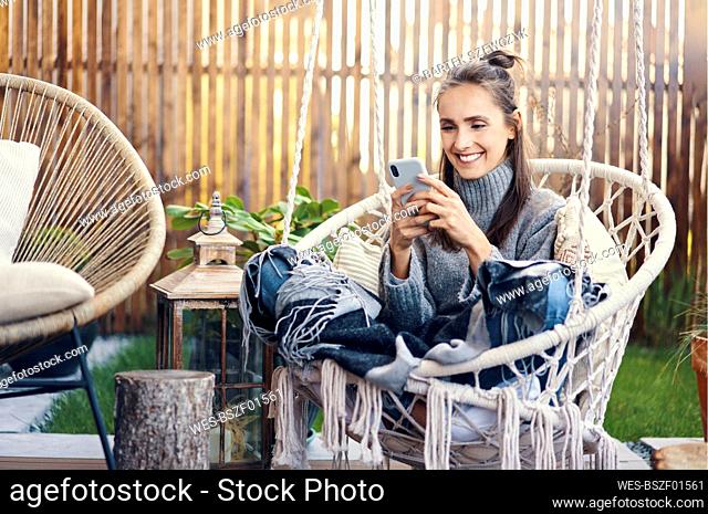 Smiling beautiful woman using smart phone while relaxing on swing in garden