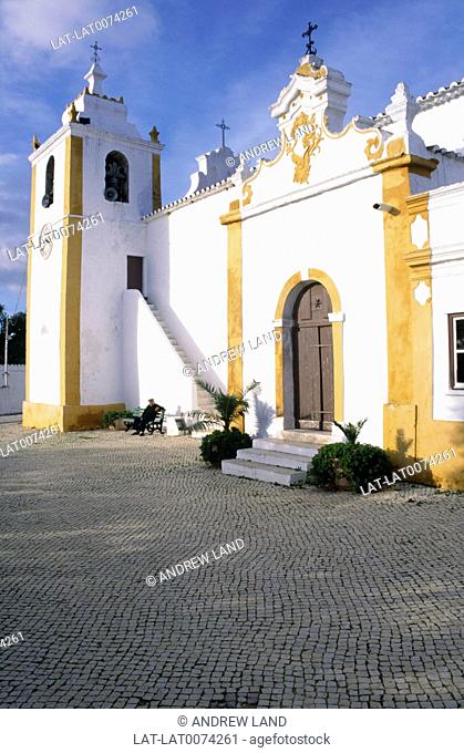 A traditional church building with white and yellow painted plaster walls with square bell tower. A man sitting on a bench