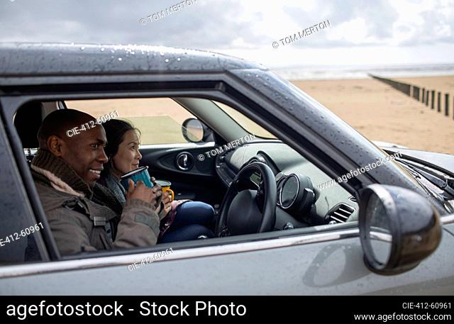 Happy couple drinking tea in car on wet winter beach