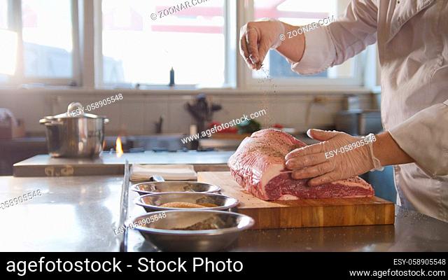 Butcher adding spices into large piece of fresh raw meat lying on a wooden board in a commercial kitchen, cooking and Haute cuisine concept