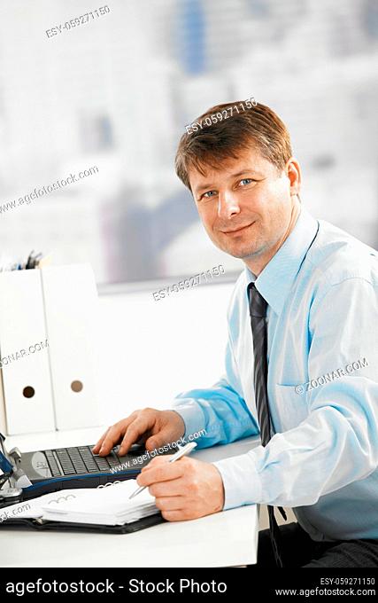 Businessman working at desk, using laptop and personal organizer. Looking at camera