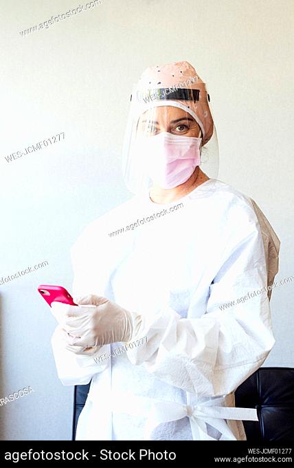 Dentist in protective suit using mobile phone while standing at office