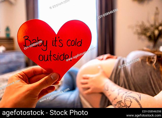 A selective focus first person perspective of a hand holding a red heart with message baby its cold outside, as heavily pregnant woman rests in bed