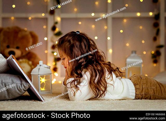 Happy curly little child girl is reading fairy tale book on the background with lights. Merry Christmas
