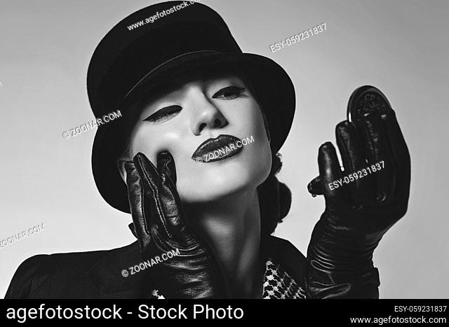 Beautiful young woman with red lips make-up in retro black and white dress, hat, jacket and leather gloves standing on grey background looking at pocket mirror