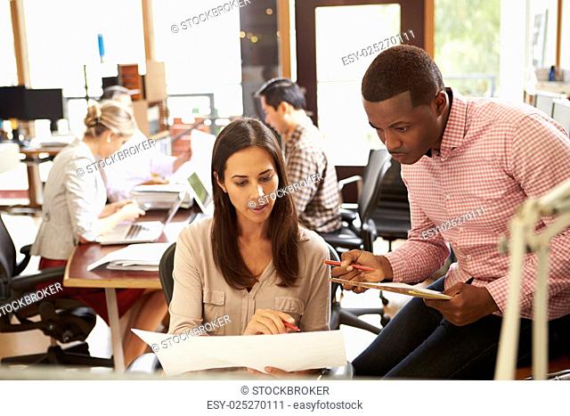 Two Colleagues Working At Desk With Meeting In Background
