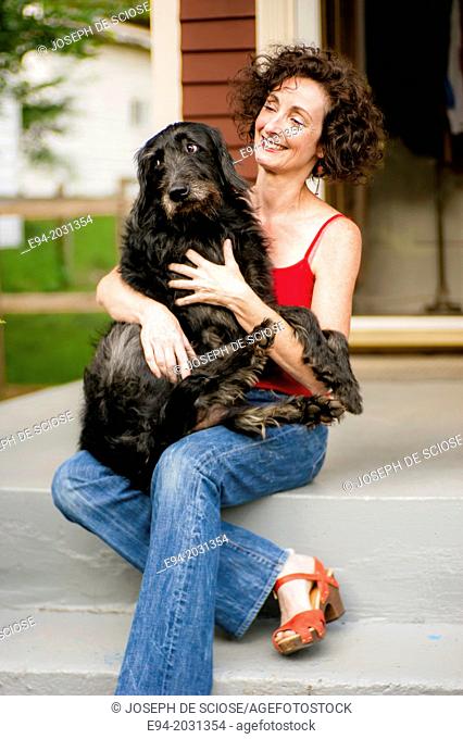 A 51 year old brunette woman wearing jeans sitting on steps in front of her house playing with her dog