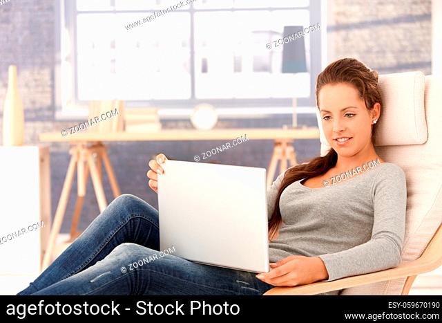Attractive young female browsing internet on laptop, sitting in armchair at home, smiling