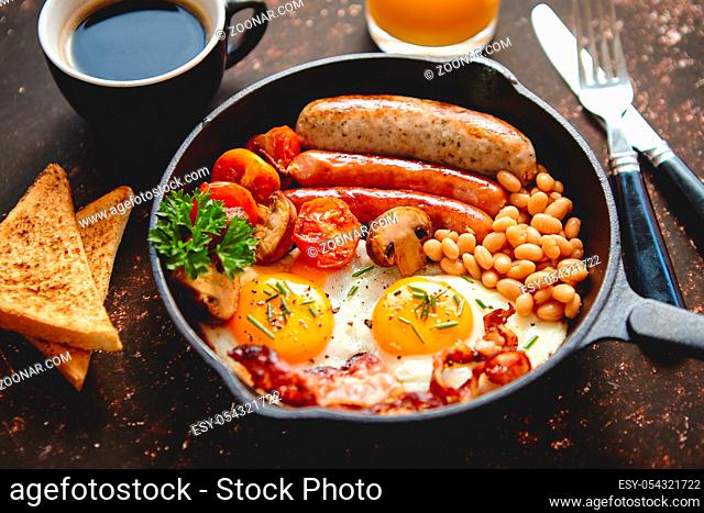 Full English breakfast - fried egg, baked beans, bacon, sausages on a dark rusty background, toasts, orange juice and coffee on a side