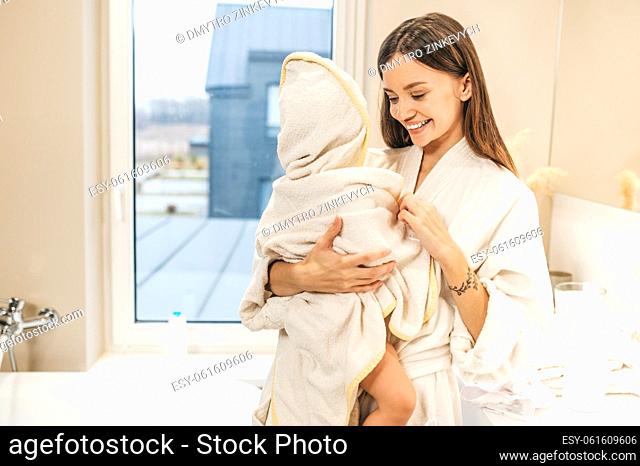 Smiling pleased beautiful young mother holding her baby dressed in a white bathrobe after bathing