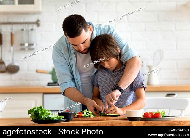 Caring father teaching cooking little son showing him how preparing vegetable healthy salad cutting cucumber on wooden board, shares knowledge, gives skills