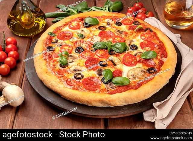 A photo of pepperoni pizza with a glass of white wine, olive oil, basil leaves, mushrooms, and cherry tomatoes, on a pizza stone, on a dark rustic background
