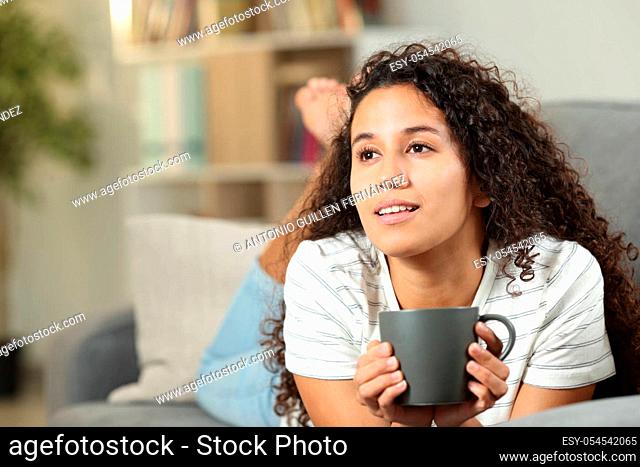 Pensive latin woman drinking coffee lying on a couch at home