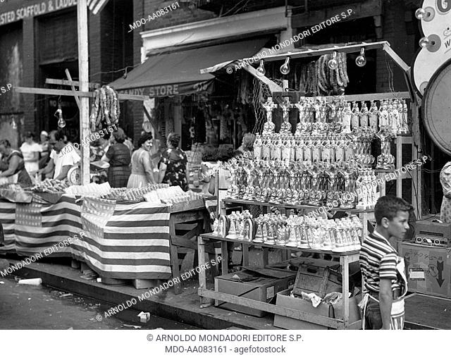 Market stalls in Little Italy. Stall selling sacred goods next to a stall selling American flags. New York, 1950s