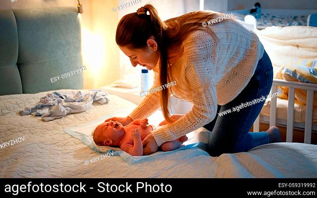 Young caring mother undressing her little baby and changing messy diapers at night before going to sleep. Concept of loving and caring parents